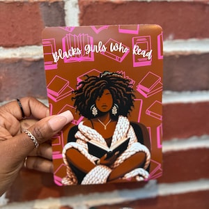 May include: A brown rectangular item with the text "Blacks girls who read" in pink. The image features a Black woman reading a book, surrounded by book illustrations. The woman is wearing a white shawl and jewelry.