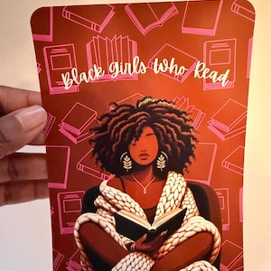 May include: A rectangular print featuring a dark-skinned person with curly hair reading a book while wrapped in a white chunky knit blanket. The background is a deep rust color with a repeating pattern of pink book outlines and the text Black girls who Read.