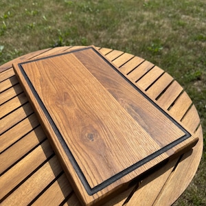 Può includere: Un tagliere rettangolare in legno caratterizzato da un centro con venature color marrone chiaro naturale e un bordo marrone scuro. Il tagliere presenta una superficie liscia e piatta con una scanalatura incassata lungo il perimetro.