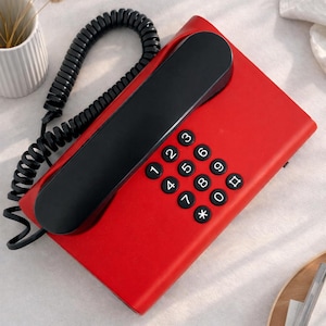 May include: A bright red telephone with a black handset and coiled cord. The keypad features white numbers and symbols. This retro-style phone would suit a modern home or office setting, adding a pop of colour.