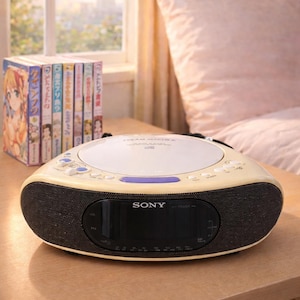 Puede incluir: Un reproductor de CD y radio Sony color crema está sobre una mesita de noche de madera. La radio tiene una rejilla de altavoz negra y una pantalla digital. Una pila de libros coloridos está a la izquierda. Una ventana y una cama están al fondo.