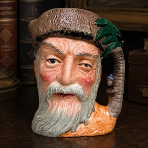 May include: A ceramic character mug shaped like a bearded man wearing a brown fur hat. The handle is designed as a palm tree with green fronds and a small figure standing at the base. The man has a grey beard, pink cheeks, and a tan-coloured base.