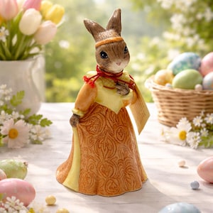 以下が含まれることがあります： 黄色とオレンジ色のドレスと赤いスカーフを着た陶器のイースターバニーの置物。バニーは、イースターエッグ、花、バスケットを背景にした白い表面に立っています。置物には茶色の耳とヘッドバンドが付いています。