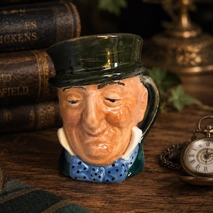 May include: A ceramic character mug shaped like a person wearing a dark green top hat, a white collared shirt, and a blue bow tie with black polka dots. The face features a tan complexion with closed eyes and a subtle smile, with a handle on the side.
