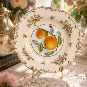 May include: A cream-coloured decorative plate with a scalloped edge and gold floral accents. The centre features a painting of two oranges on a branch. The plate is displayed on a gold-coloured metal stand, showcasing a vintage aesthetic.