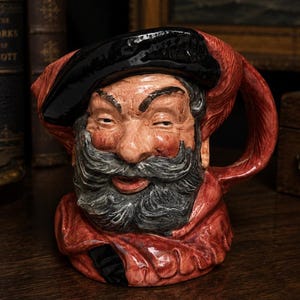 Puede incluir: Una jarra de cerámica con forma de personaje con una espesa barba y bigote grises, que lleva una boina negra y una prenda roja. La figura tiene rasgos faciales detallados con textura de piel visible, pintada en tonos rojos, grises y negros.