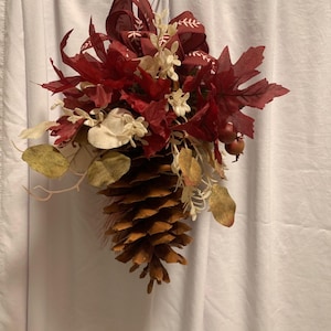 May include: A hanging decorative ornament featuring a large brown pinecone at the base, topped with deep red maple leaves, cream-colored sprigs, small round berries, and a dark red ribbon with a white leaf pattern tied in a bow at the top.