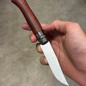 May include: A folding knife with a wooden handle and a stainless steel blade. The handle is a dark reddish-brown color, and the blade is silver. The knife is held in a hand.