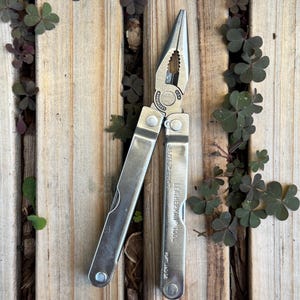 May include: A silver Leatherman multi-tool with needle-nose pliers, wire cutters, and folding handles. The metal body features engraved text reading Leatherman Tool, U.S. Reg. TM 1325472, and Portland OR. The tool is shown in an open position.