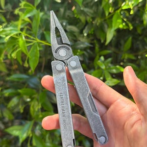 May include: A silver multi-tool with pliers, a knife, and other tools. The tool is held open, displaying the pliers. The text "LEATHERMAN BOND" is visible on the handle. The background is a blurred green leafy plant.