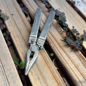 May include: A silver-colored stainless steel multi-tool featuring needle-nose pliers with a textured gripping surface. The tool has two folding handles with visible metal hinges and the Leatherman brand name engraved on the central pivot point.