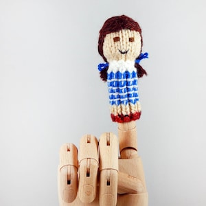 May include: A finger puppet of a girl with brown hair, wearing a blue and white gingham dress. The puppet is made of yarn and has a smiling face.