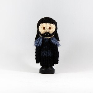 May include: A crocheted doll with black hair, a black beard, and a black and blue outfit. The doll is standing on a wooden base.