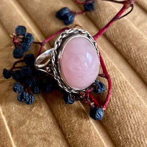 May include: A silver ring featuring a large, oval, pale pink gemstone. The gemstone is set in a silver bezel with a rope-like design. The ring is displayed with dark blue berries and a red stem.