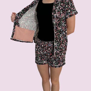 May include: Black floral print pajama set with a short-sleeved button-up shirt, shorts, and a black tank top. The set features a white trim and a pink floral pattern. The shirt is open, revealing a pink rectangular object in the pocket.