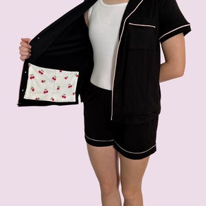 May include: Black pajama set with white piping trim. The short-sleeved top has a chest pocket and is open, revealing a cherry-printed pocket lining. The matching shorts also have white trim. A white tank top is worn underneath.