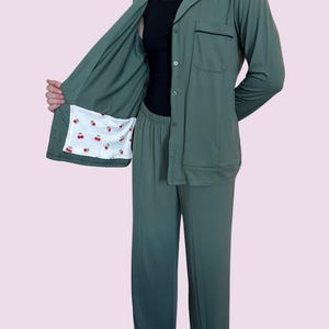 May include: Sage green pajama set. The long-sleeved top has a notched collar, a chest pocket, and black trim. The inside lining of the jacket features a white fabric with a red cherry pattern. The set includes matching pants.