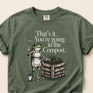 May include: Moss green t-shirt with a humorous graphic of a frog in an apron holding a pitchfork, standing next to a compost bin. Text reads "That's it. You're going in the Compost."