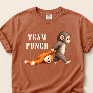 May include: A rust-colored t-shirt with rolled-up sleeves. The shirt features the text "TEAM PUNCH" in white block letters. An illustration depicts a gray monkey pulling an orange monkey by the arm.