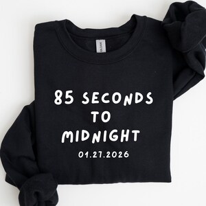 May include: Black sweatshirt with white text that reads "85 SECONDS TO MIDNIGHT" and the date "01.27.2026". The sweatshirt is folded and has a crew neck.