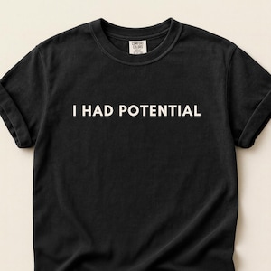 May include: Black t-shirt with the text "I HAD POTENTIAL" in white. The shirt has rolled-up sleeves and a Comfort Colors tag. The bottom left corner of the image has the word "BLACK" in a black rectangle. The bottom of the image has text.