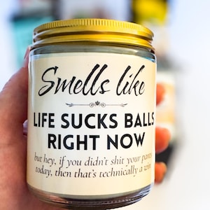 Op de afbeelding: Een doorzichtige glazen kaarsenpot met een goudkleurig metalen deksel. Het etiket bevat de zwarte tekst: Smells like LIFE SUCKS BALLS RIGHT NOW but hey, if you didn't shit your pants today, then that's technically a win. De kaarswas is zichtbaar.