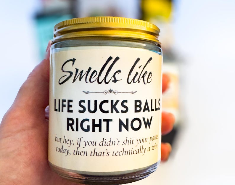 Op de afbeelding: Een doorzichtige glazen kaarsenpot met een goudkleurig metalen deksel. Het etiket bevat de zwarte tekst: Smells like LIFE SUCKS BALLS RIGHT NOW but hey, if you didn't shit your pants today, then that's technically a win. De kaarswas is zichtbaar.