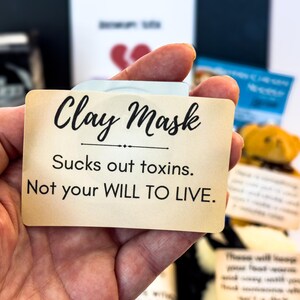 Puede incluir: Una tarjeta beige con las palabras "Clay Mask" en escritura negra, con el texto "Sucks out toxins. Not your WILL TO LIVE." La tarjeta se sostiene en una mano, con otros elementos en el fondo.