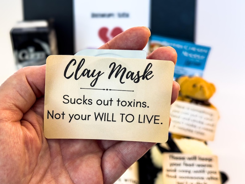 Puede incluir: Una tarjeta beige con las palabras "Clay Mask" en escritura negra, con el texto "Sucks out toxins. Not your WILL TO LIVE." La tarjeta se sostiene en una mano, con otros elementos en el fondo.