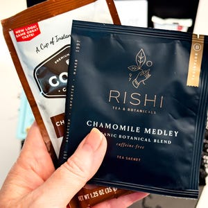 Op de afbeelding: Een donkerblauw Rishi Tea & Botanicals Chamomile Medley theezakje met gouden tekst en een handicoon, vastgehouden voor een bruin koffiezakje met de tekst "New Look! Same Great Taste!" en "Net Wt 1.25 oz (35 g)" onderaan.