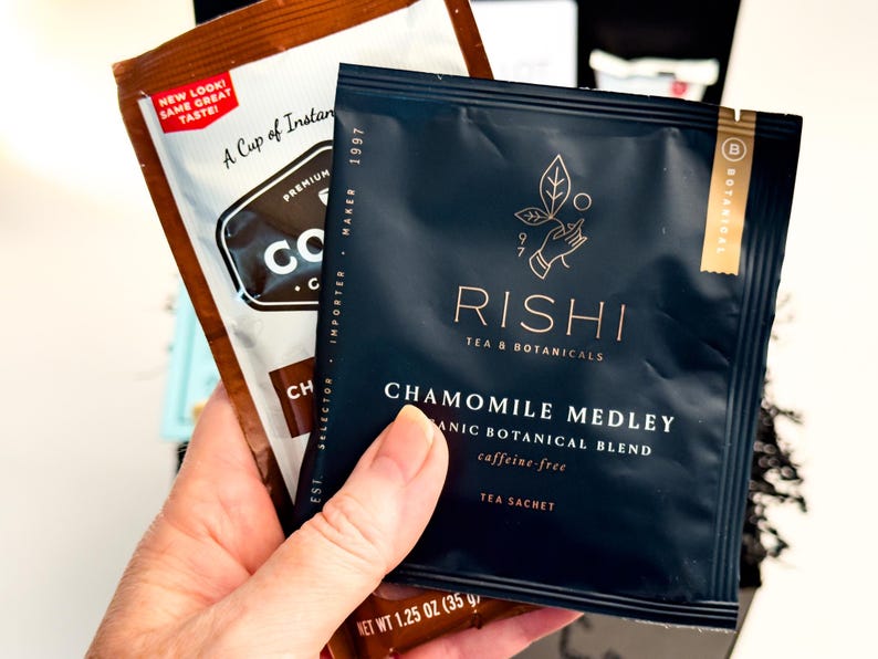 Op de afbeelding: Een donkerblauw Rishi Tea & Botanicals Chamomile Medley theezakje met gouden tekst en een handicoon, vastgehouden voor een bruin koffiezakje met de tekst "New Look! Same Great Taste!" en "Net Wt 1.25 oz (35 g)" onderaan.