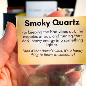 Op de afbeelding: Een doorzichtig plastic zakje met twee gladde, donkergekleurde rookkwartsstenen. Bevestigd is een rechthoekige beige kaart met zwarte tekst: Smoky Quartz, For keeping the bad vibes out, the assholes at bay, and turning that dark, heavy energy into something lighter. (And if that doesn't work, it's a handy thing to throw at someone).