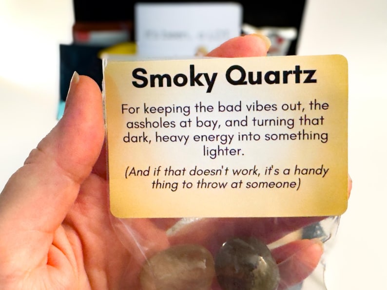 Op de afbeelding: Een doorzichtig plastic zakje met twee gladde, donkergekleurde rookkwartsstenen. Bevestigd is een rechthoekige beige kaart met zwarte tekst: Smoky Quartz, For keeping the bad vibes out, the assholes at bay, and turning that dark, heavy energy into something lighter. (And if that doesn't work, it's a handy thing to throw at someone).