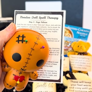 Puede incluir: Una mu&ntilde;eca vud&uacute; naranja con ojos de bot&oacute;n negros, detalles cosidos y alfileres. La mu&ntilde;eca se sostiene junto a una tarjeta con el texto "Voodoo Doll Spell Therapy". Tambi&eacute;n se ve un peque&ntilde;o osito de peluche amarillo.