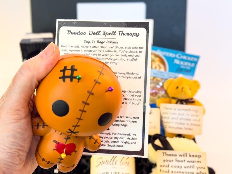 Puede incluir: Una mu&ntilde;eca vud&uacute; naranja con ojos de bot&oacute;n negros, detalles cosidos y alfileres. La mu&ntilde;eca se sostiene junto a una tarjeta con el texto "Voodoo Doll Spell Therapy". Tambi&eacute;n se ve un peque&ntilde;o osito de peluche amarillo.