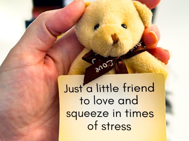 Op de afbeelding: Een kleine beige pluche teddybeer met een bruin lint om de nek met het woord love erop. De beer houdt een rechthoekige gele kaart vast met de zwarte tekst: Just a little friend to love and squeeze in times of stress.