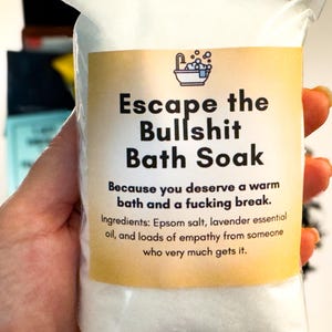 Op de afbeelding: Een doorzichtig plastic zakje gevuld met wit badzout. Het etiket bevat een icoon van een badkuip en de tekst: Escape the Bullshit Bath Soak. Because you deserve a warm bath and a fucking break. Ingredients: Epsom salt, lavender essential oil, and loads of empathy from someone who very much gets it.