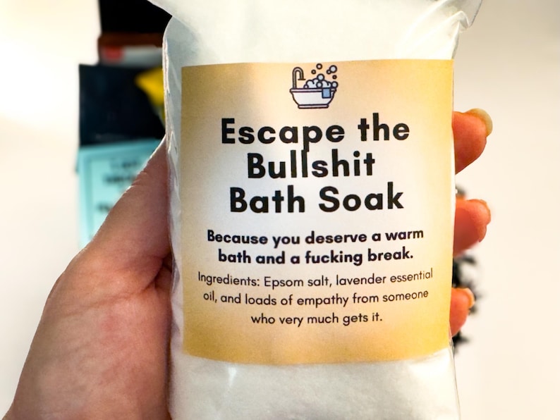 Op de afbeelding: Een doorzichtig plastic zakje gevuld met wit badzout. Het etiket bevat een icoon van een badkuip en de tekst: Escape the Bullshit Bath Soak. Because you deserve a warm bath and a fucking break. Ingredients: Epsom salt, lavender essential oil, and loads of empathy from someone who very much gets it.