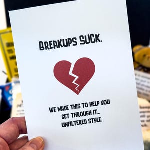 Puede incluir: Una tarjeta blanca con el texto "BREAKUPS SUCK" y un coraz&oacute;n rojo roto. La tarjeta tambi&eacute;n dice "WE MADE THIS TO HELP YOU GET THROUGH IT... UNFILTERED STYLE."