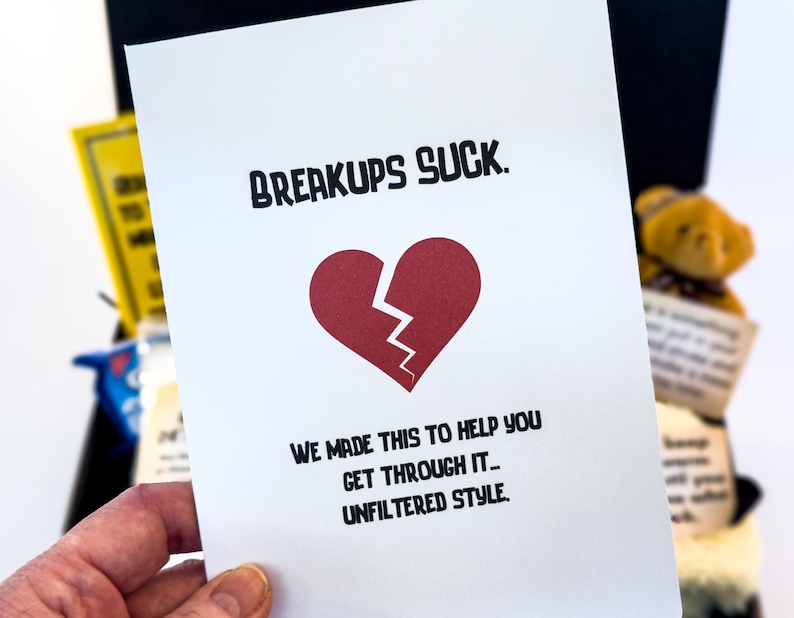 Puede incluir: Una tarjeta blanca con el texto "BREAKUPS SUCK" y un coraz&oacute;n rojo roto. La tarjeta tambi&eacute;n dice "WE MADE THIS TO HELP YOU GET THROUGH IT... UNFILTERED STYLE."