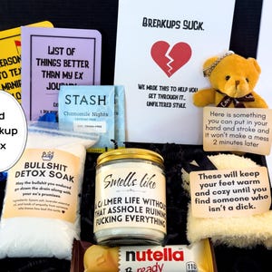 Puede incluir: Una caja negra llena de art&iacute;culos de recuperaci&oacute;n de rupturas. Incluye un diario, t&eacute;, sales de ba&ntilde;o, una vela, un osito de peluche y un snack de Nutella. La caja tiene el texto "Bad Breakup Box".