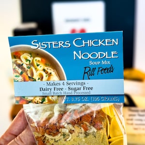Puede incluir: Un paquete de mezcla para sopa de fideos con pollo Sisters. La etiqueta azul muestra el nombre del producto, una imagen de sopa y el texto "Makes 4 Servings - Dairy Free - Sugar Free". Peso neto: 106 gramos.