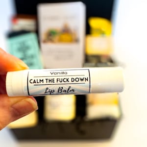 Op de afbeelding: Een witte cilindrische lippenbalsemhuls met een etiket met de tekst Vanilla, CALM THE FUCK DOWN en Lip Balm in zwarte letters. Het etiketontwerp bevat lichtblauwe zonnestralen op een witte achtergrond met een dunne zwarte rand.