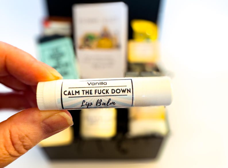 Op de afbeelding: Een witte cilindrische lippenbalsemhuls met een etiket met de tekst Vanilla, CALM THE FUCK DOWN en Lip Balm in zwarte letters. Het etiketontwerp bevat lichtblauwe zonnestralen op een witte achtergrond met een dunne zwarte rand.