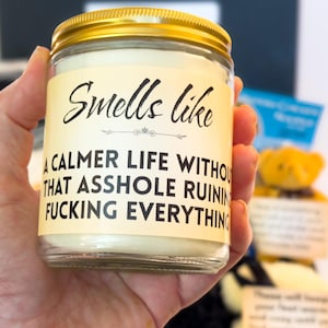Puede incluir: Una vela en tarro de cristal transparente con tapa dorada. La etiqueta dice: "Smells like a calmer life without that asshole ruining fucking everything."