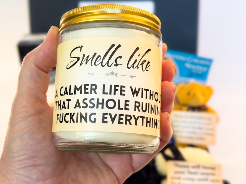 Puede incluir: Una vela en tarro de cristal transparente con tapa dorada. La etiqueta dice: "Smells like a calmer life without that asshole ruining fucking everything."
