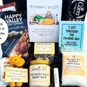 Op de afbeelding: Een geschenkdoos met Happy Valley soepmix, Carr's crackers, Smoked Gouda kaasspread, een gele teddybeer, een kaars met het label Smells like LIFE SUCKS BALLS RIGHT NOW, lippenbalsem en een zakje Escape the Bullshit badzout.