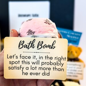 Puede incluir: Una bomba de ba&ntilde;o rosa se sostiene frente a un letrero que dice "Bath Bomb". El letrero contin&uacute;a con texto humor&iacute;stico. Un peque&ntilde;o oso de peluche es visible en el fondo.