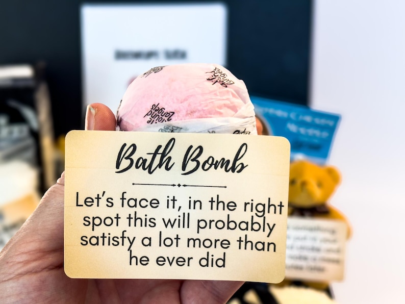 Puede incluir: Una bomba de ba&ntilde;o rosa se sostiene frente a un letrero que dice "Bath Bomb". El letrero contin&uacute;a con texto humor&iacute;stico. Un peque&ntilde;o oso de peluche es visible en el fondo.