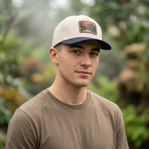 Peut inclure: Une casquette de baseball avec une calotte couleur crème et une visière bleu marine foncé. L'avant de la casquette présente un écusson rectangulaire affichant une illustration de paysage avec un lac, des montagnes et des pins dans des tons terreux.